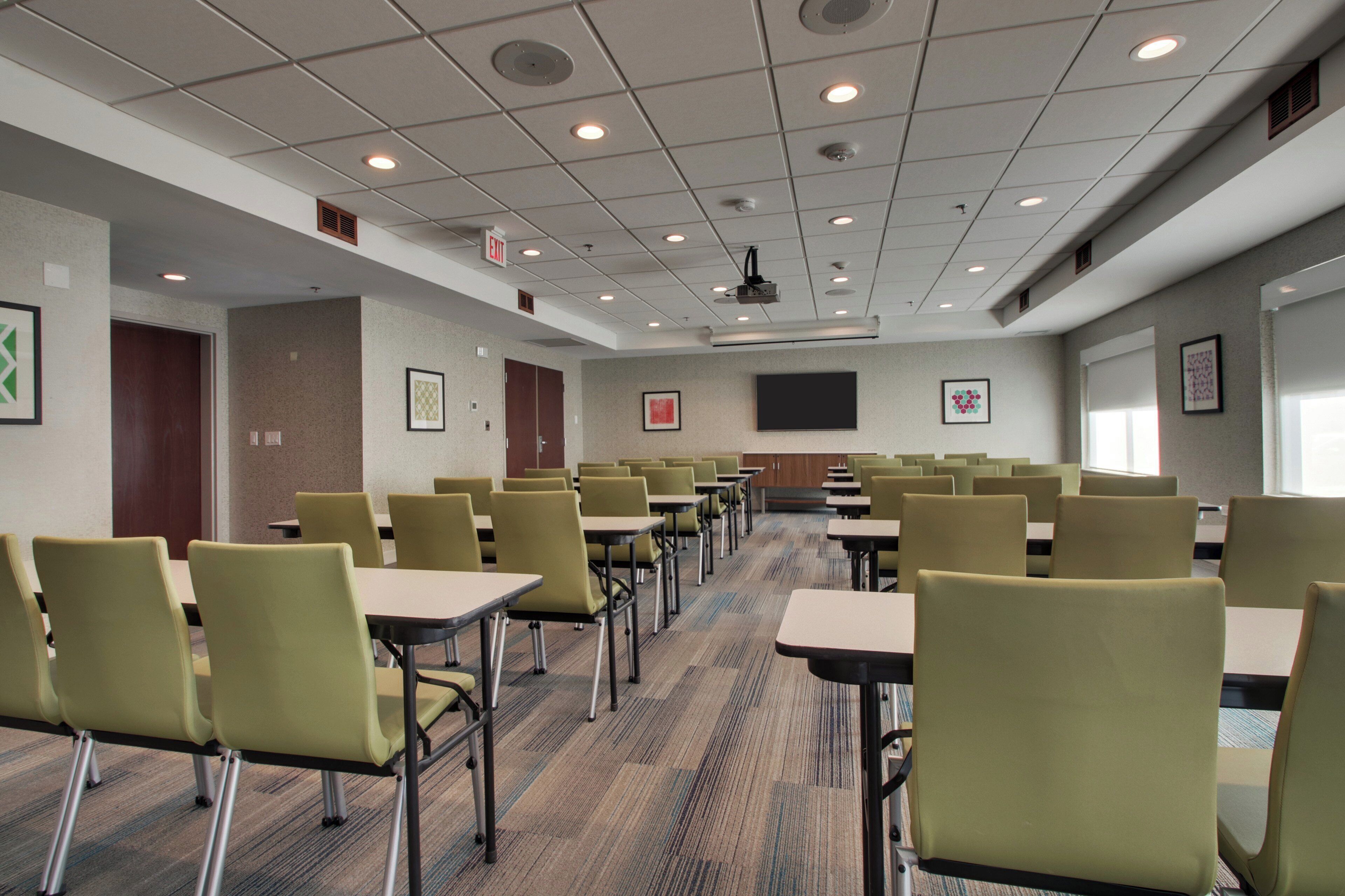 A conference room with rows of tables and chairs, a projector screen, and an exit sign.