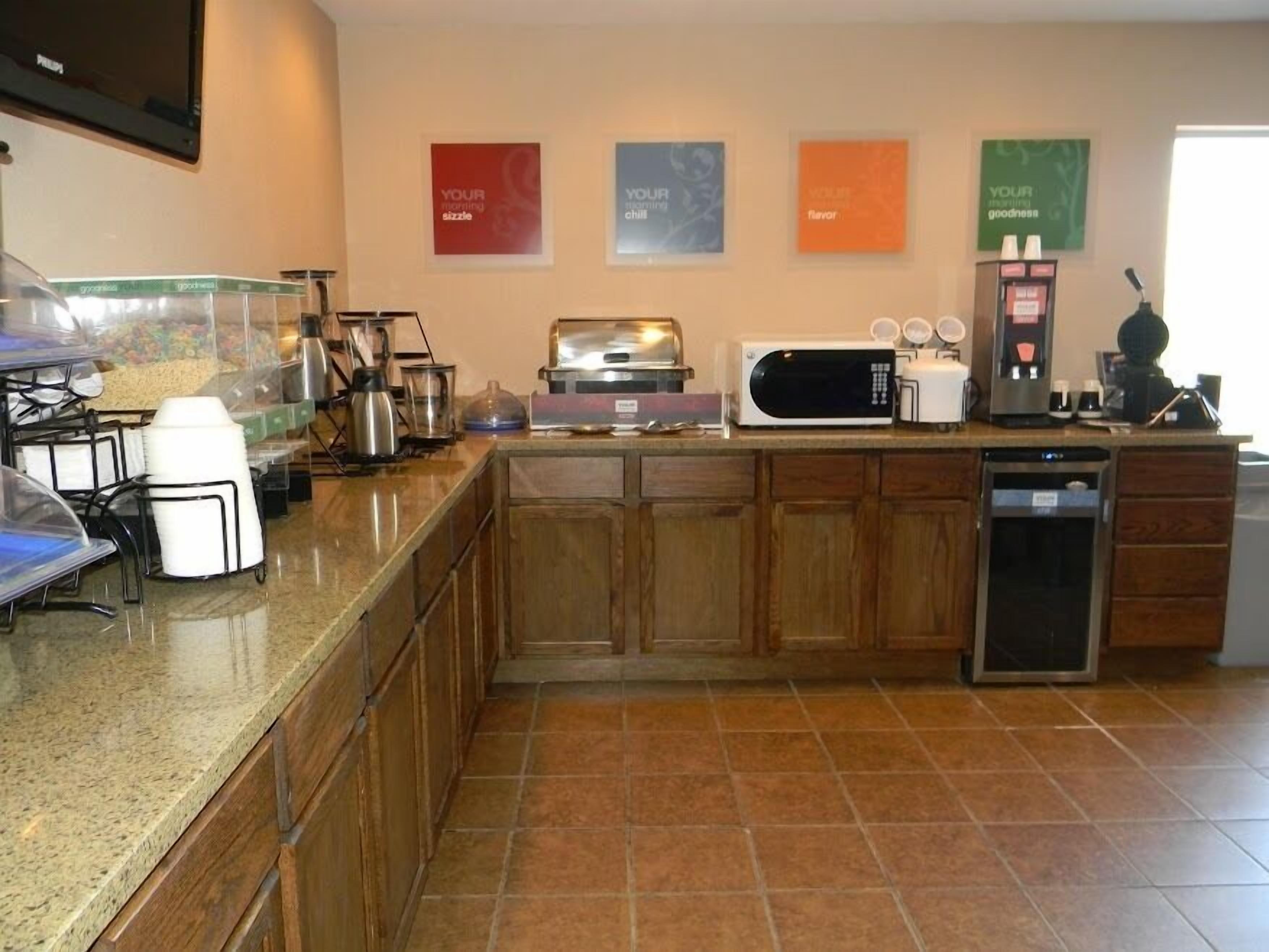 Una cocina con microondas, tostadora, cafetera y dispensador de café. Cuatro letreros enmarcados adornan la pared, cada uno con un mensaje diferente.