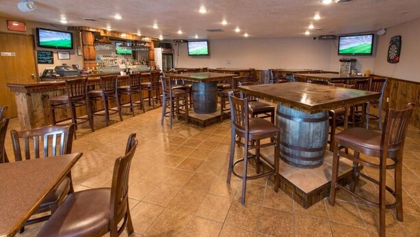 Un área de bar con mesas y sillas de madera, una barra y dos televisores montados en la pared.