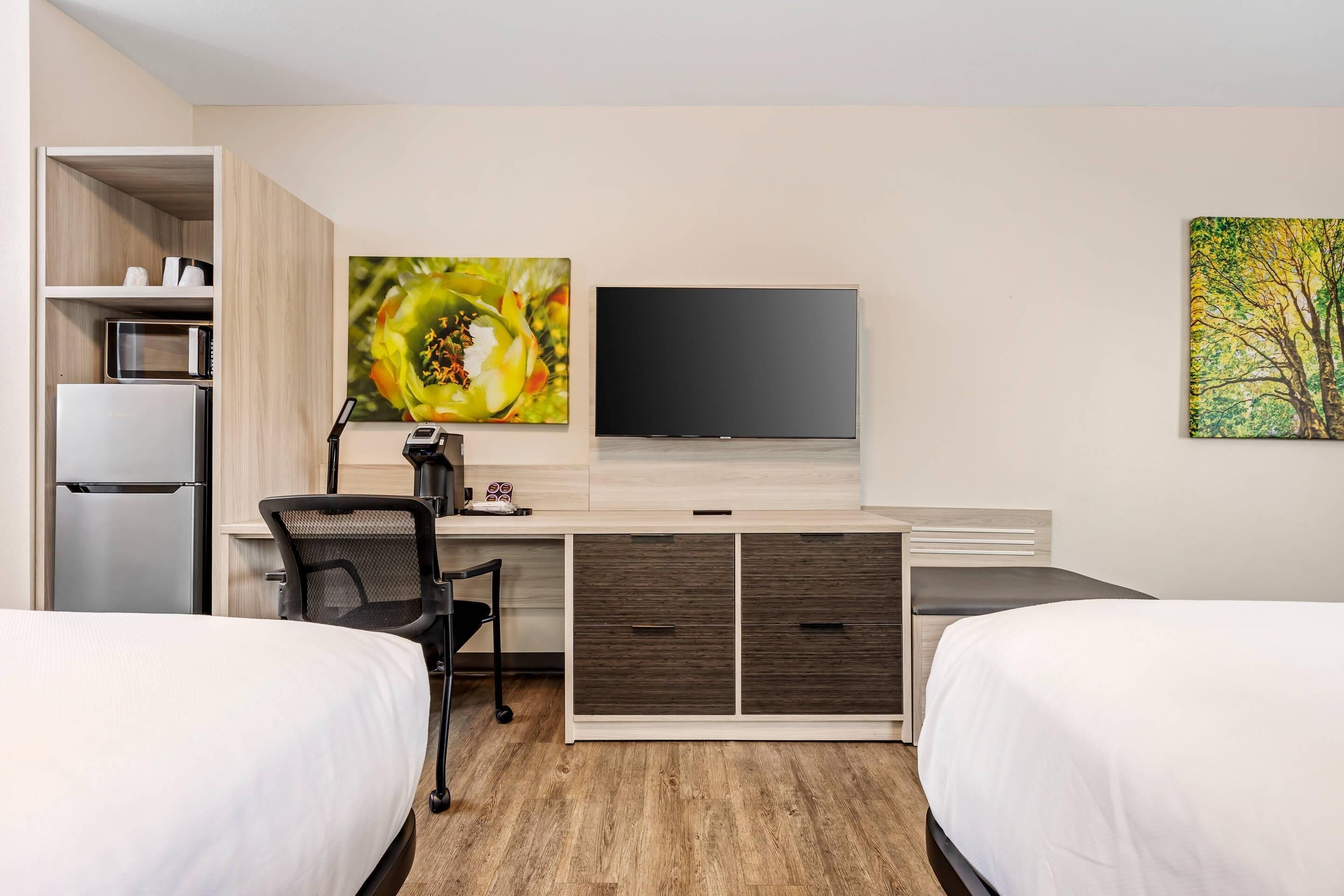 A hotel room with two beds, a desk with a chair, a flat-screen TV, a microwave, and a refrigerator.