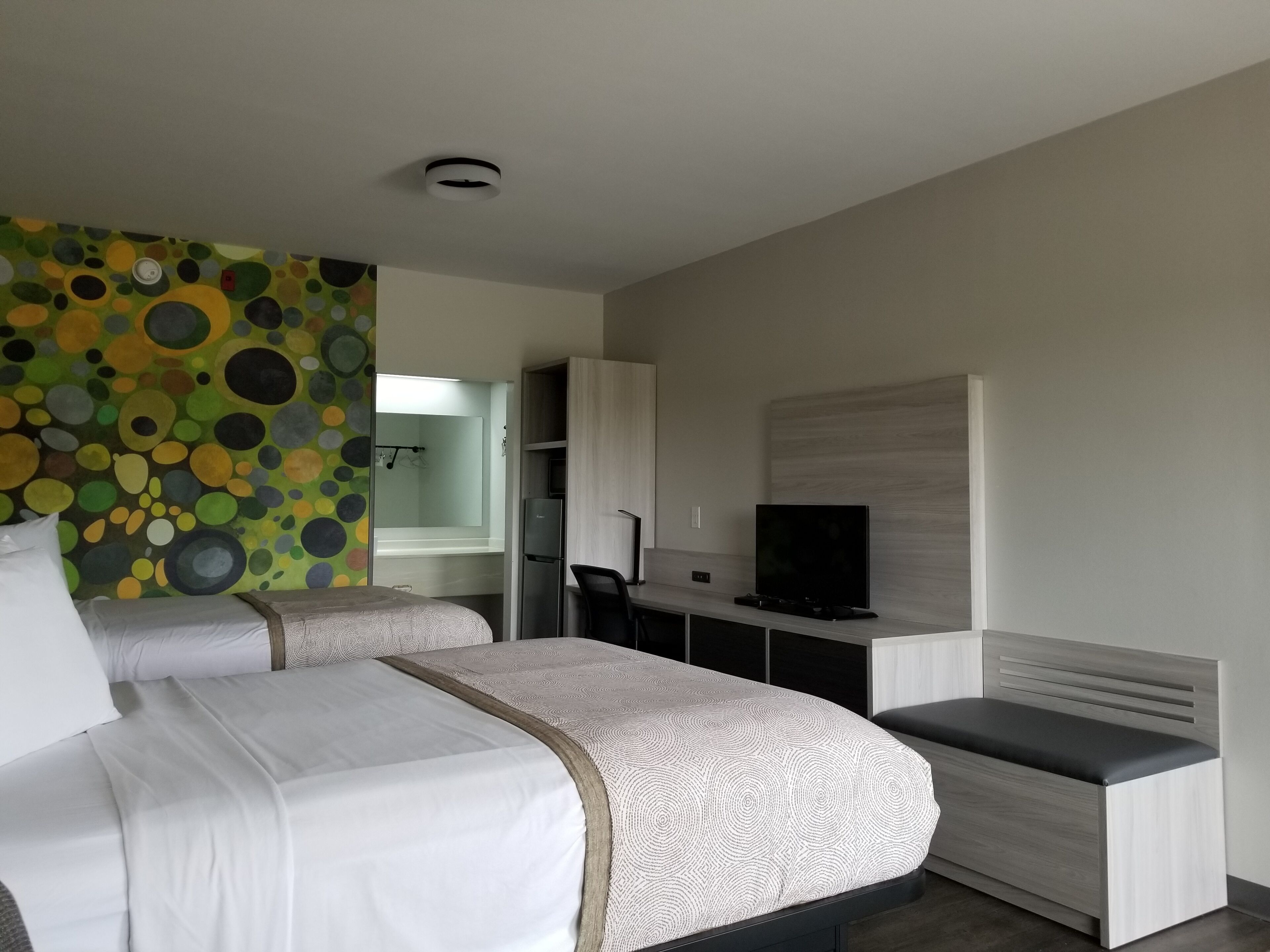 A modern hotel room with two beds, a television, a desk, and a bathroom visible through an open door.