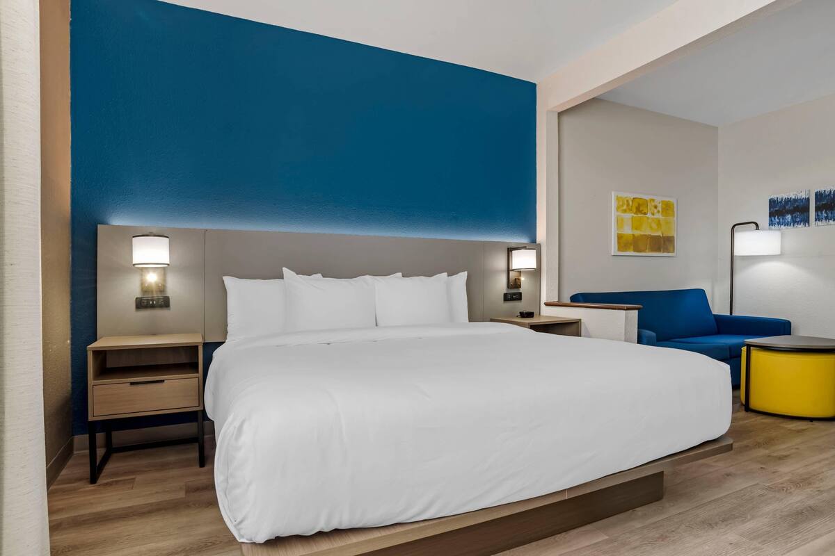 A modern hotel room with a large bed, bedside tables with lamps, a blue accent wall, and a small seating area with a yellow side table.