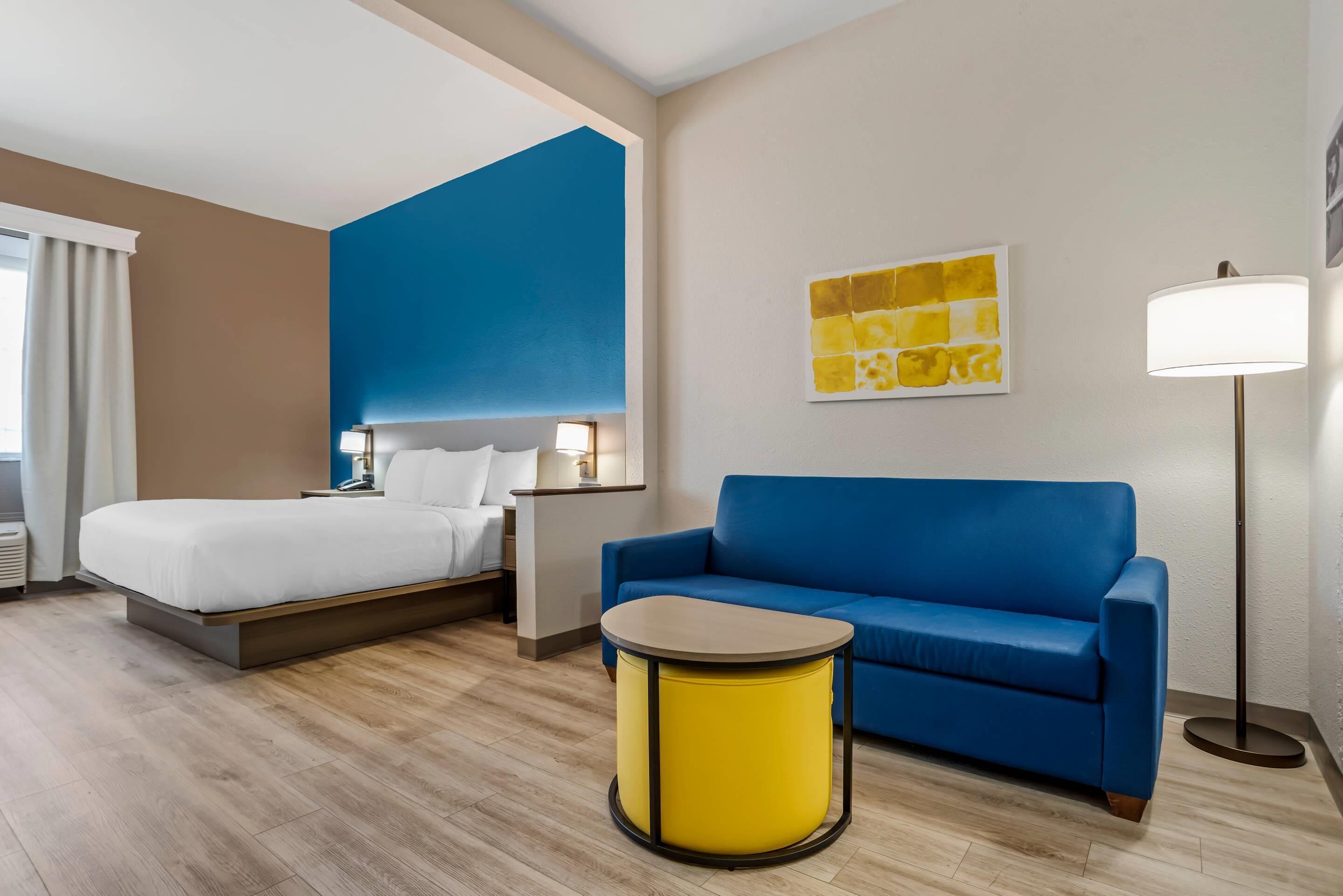Una habitación de hotel con una cama, un sofá azul, una mesita amarilla y una lámpara de pie.