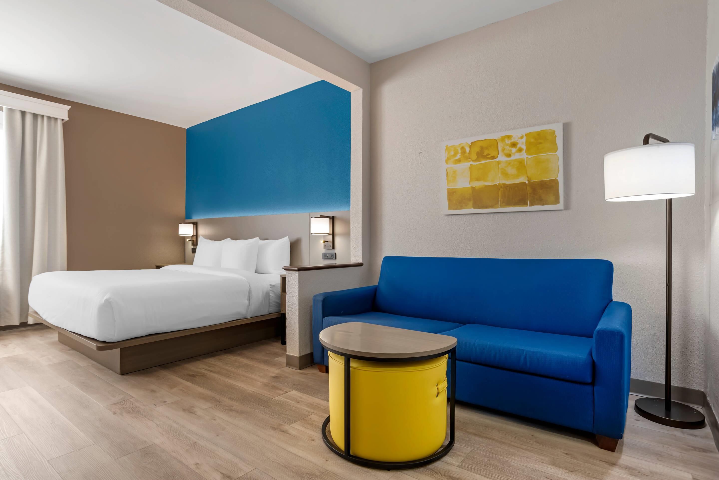 A hotel room with a bed, a blue sofa, a yellow side table, and a painting on the wall.