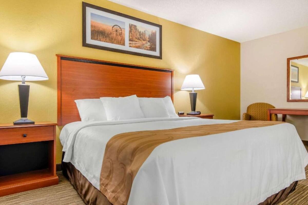 Quarto de hotel com uma cama grande, duas mesas de cabeceira com luminárias, uma cabeceira de madeira e um quadro emoldurado acima da cama.