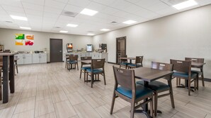 Un grand espace à manger avec des tables et des chaises en bois, une cuisine à l’arrière-plan et un sol carrelé et impeccable.