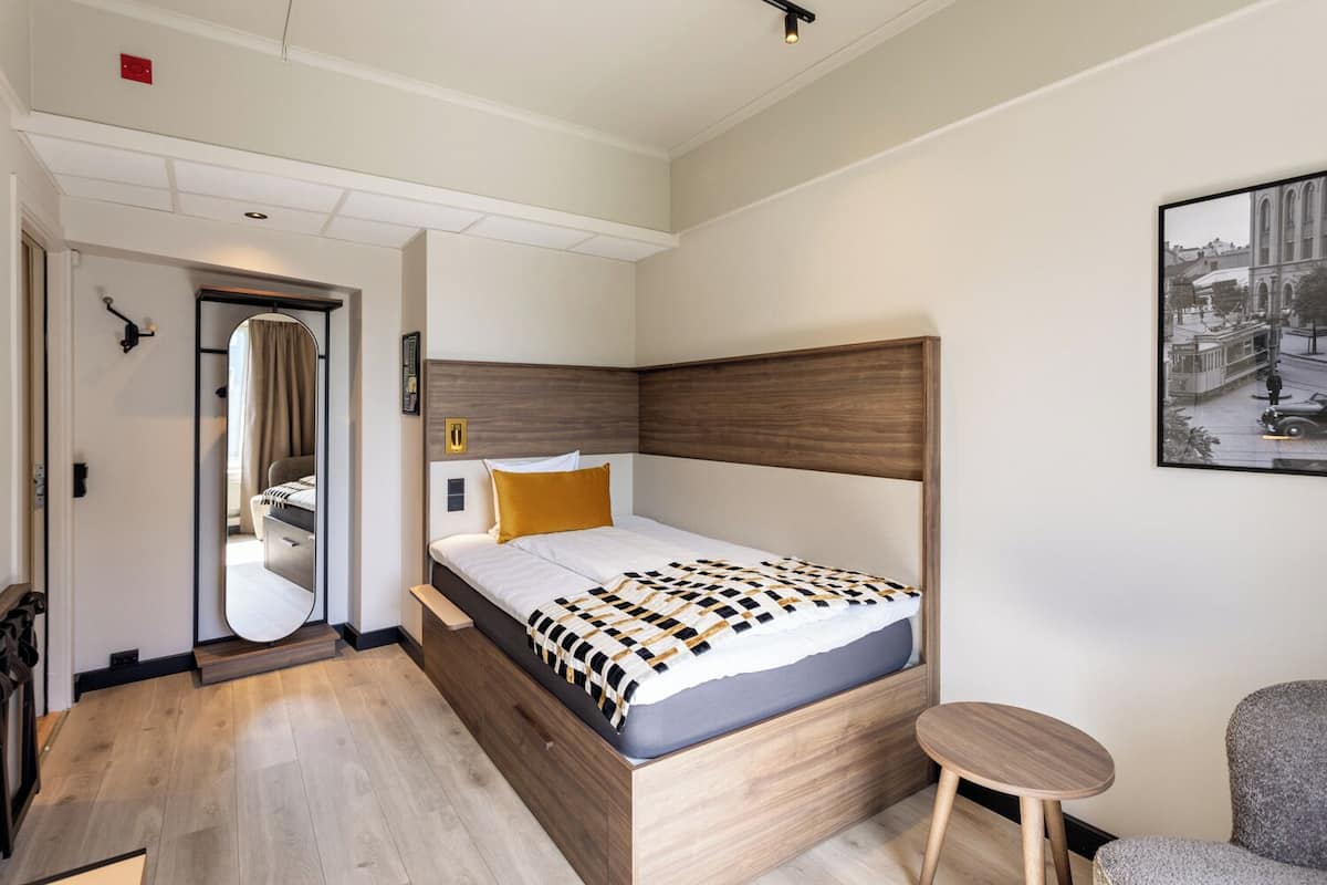 Quarto de hotel com cama de madeira, uma pequena mesa redonda, um espelho e um quadro na parede.