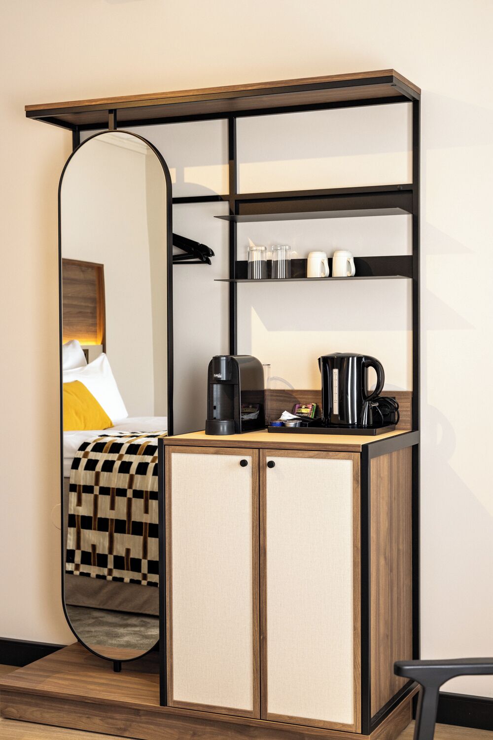 A compact hotel room with a mirrored wardrobe, a bed with a checkered blanket, and a coffee station with a kettle and coffee maker.