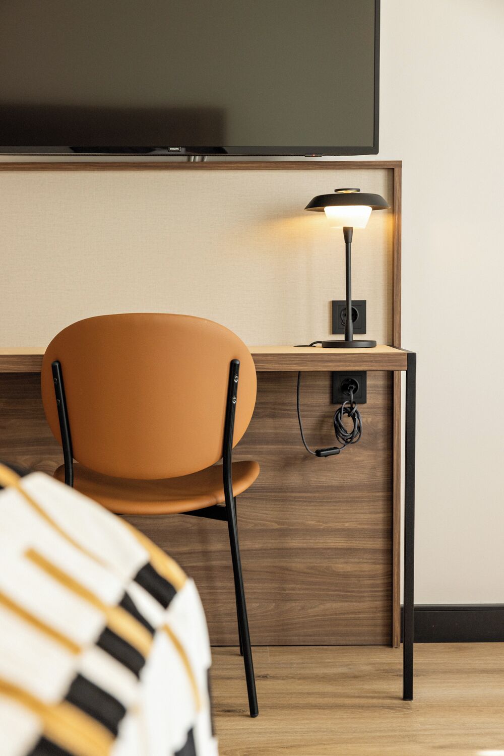 Een hotelkamer met een houten bureau, een stoel, een aan de muur bevestigde televisie en een lamp op het bureau.