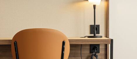 A hotel room with a wooden desk, a chair, a TV mounted on the wall, and a lamp on the desk.