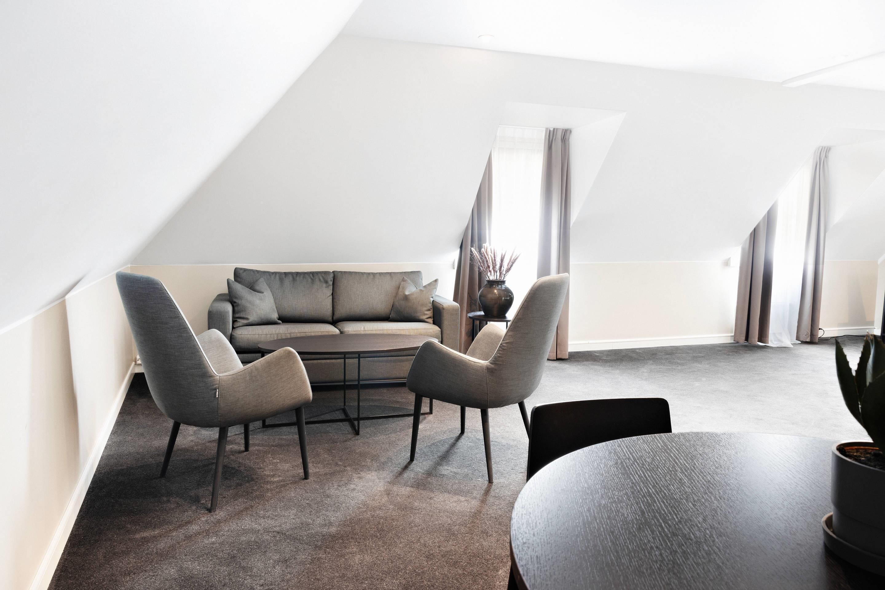 Un dormitorio estilo loft moderno con techo inclinado, un sofá gris, dos sillones, una mesita redonda y una planta en maceta.
