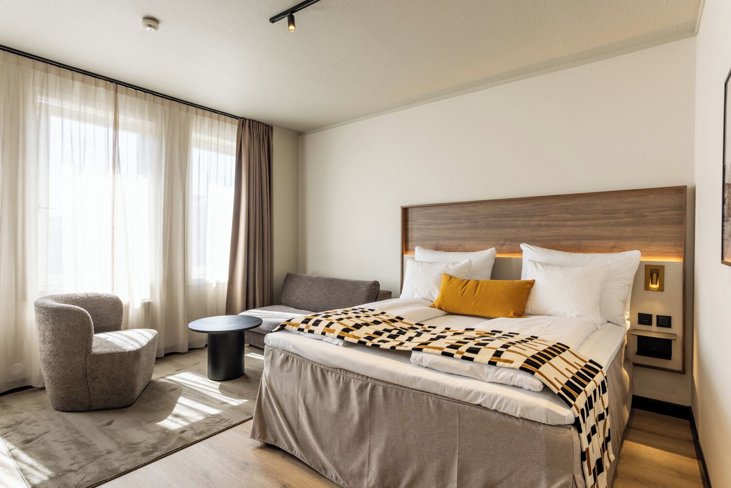 A modern hotel room with a large bed, a sofa, a chair, a small table, and a nightstand with a lamp.