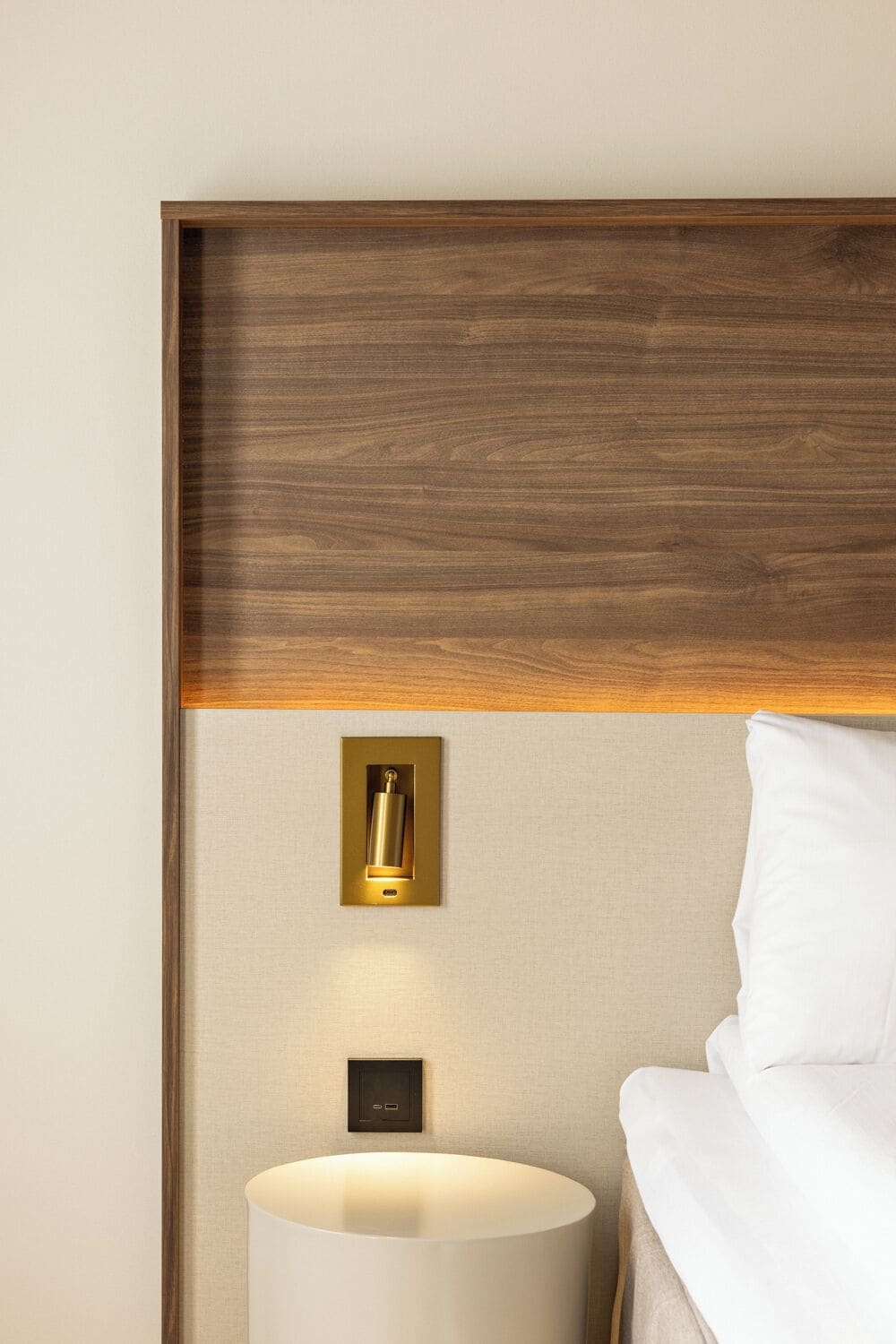 A hotel room with a wooden headboard, a bedside lamp, a wall-mounted light switch, and a white bed.