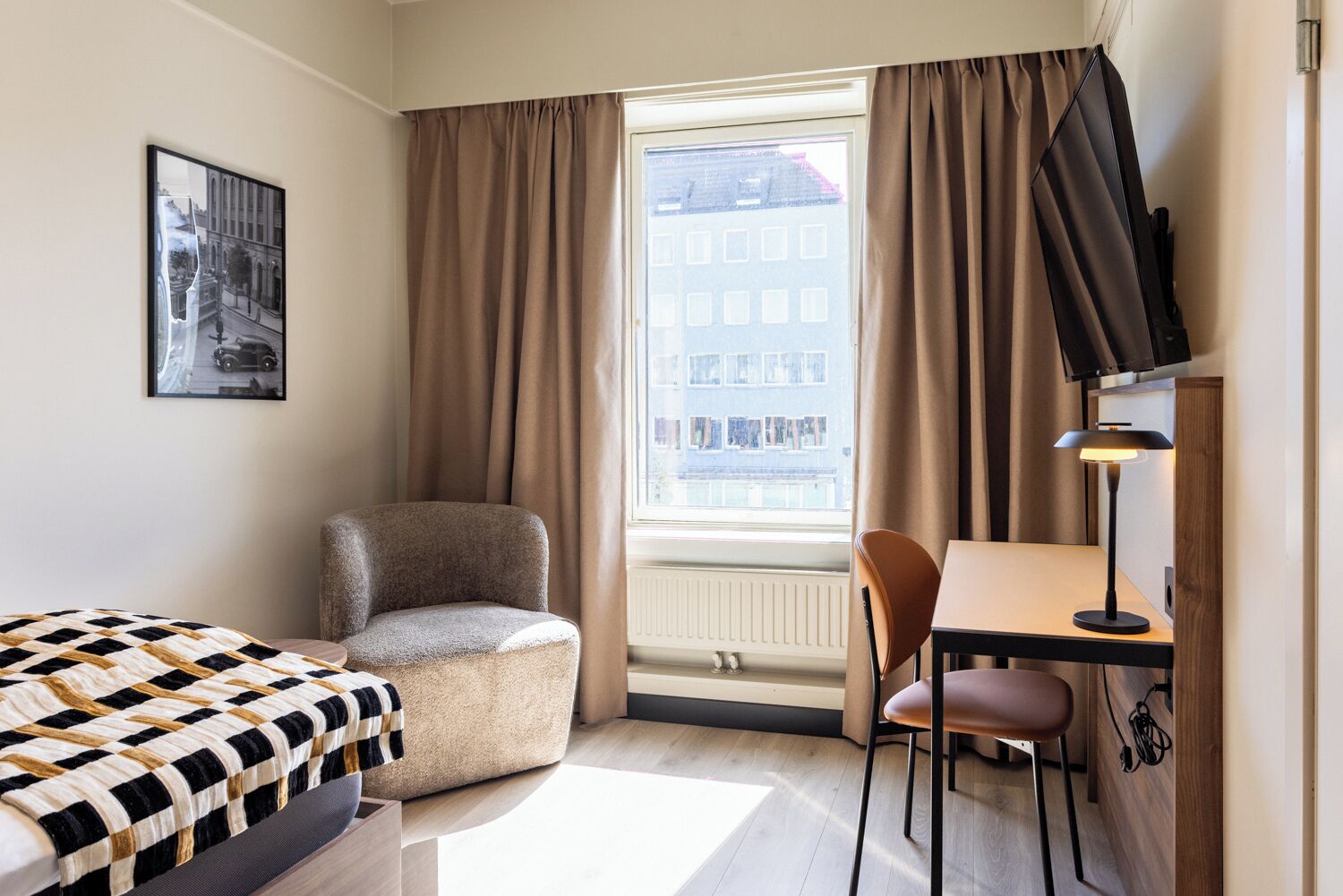 A hotel room with a bed, a chair, a desk, a lamp, a television, and a window with curtains.