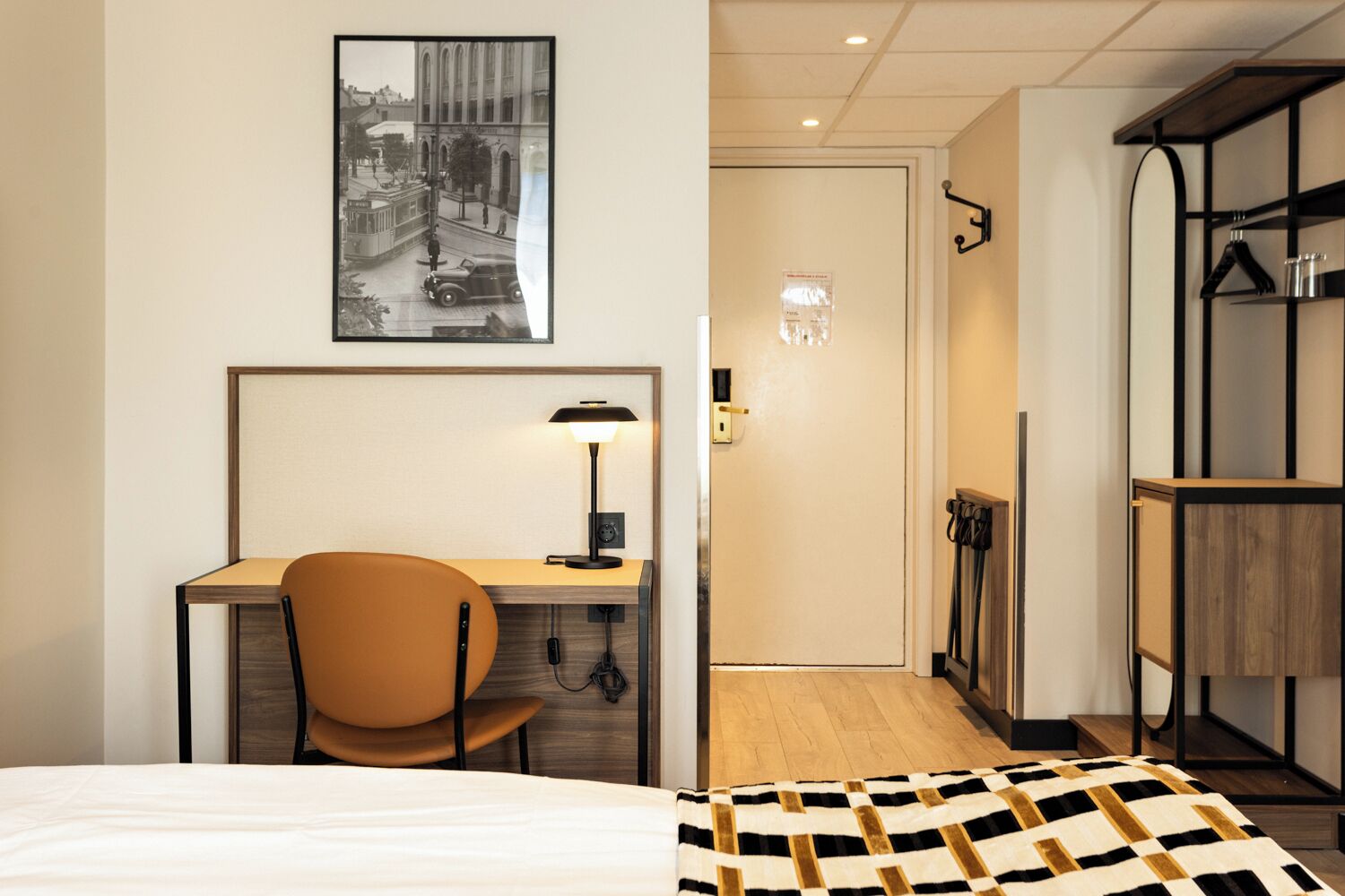 Habitación de hotel con cama, escritorio con silla, lámpara, un cuadro en la pared y piso de madera.