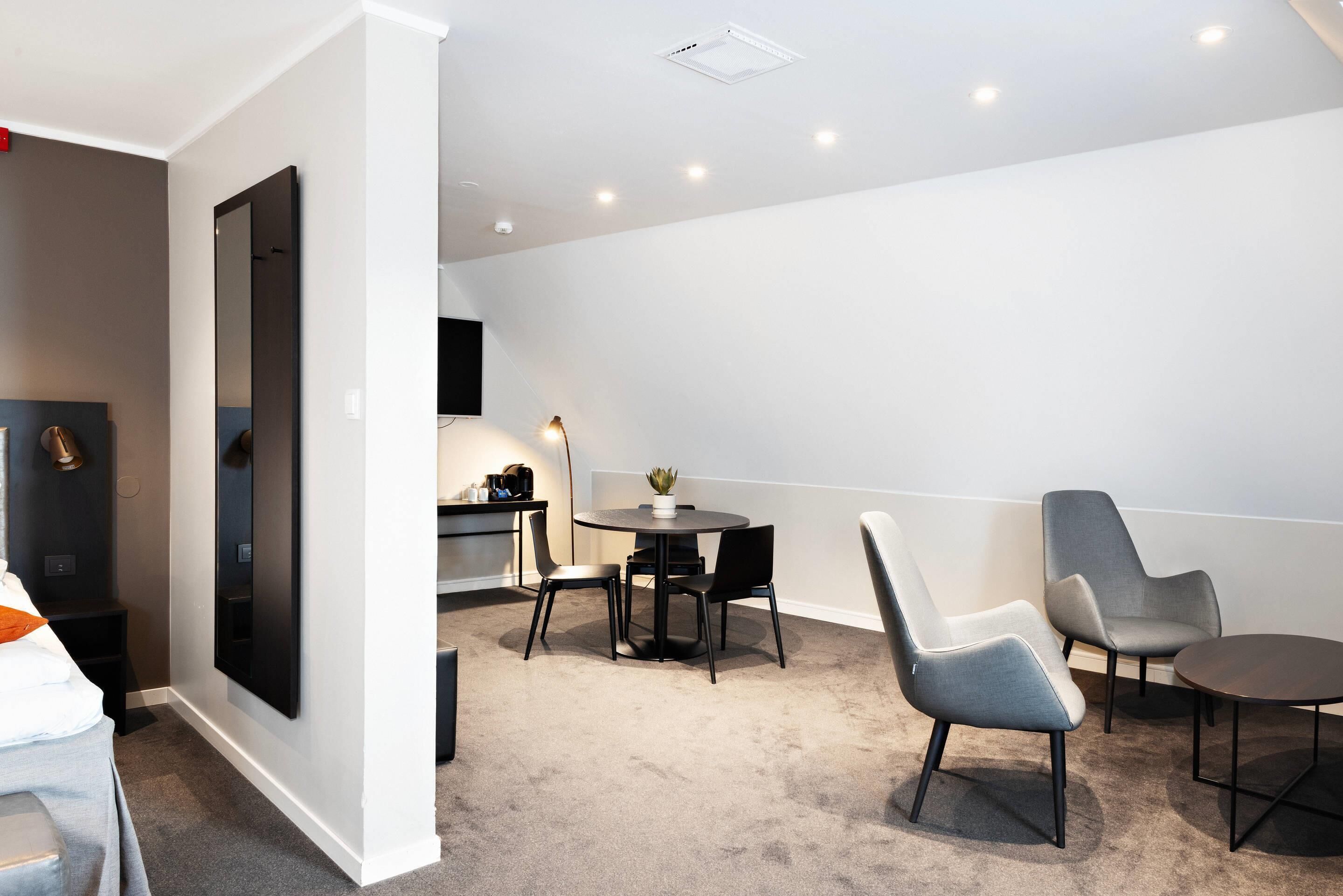 A modern hotel room with a large bed, a desk, a dining table with chairs, and a TV mounted on the wall.