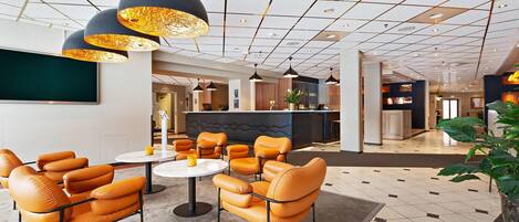 Un lobby moderno de hotel con una zona de estar que incluye mesas redondas y sillas naranjas, un bar y una planta grande.