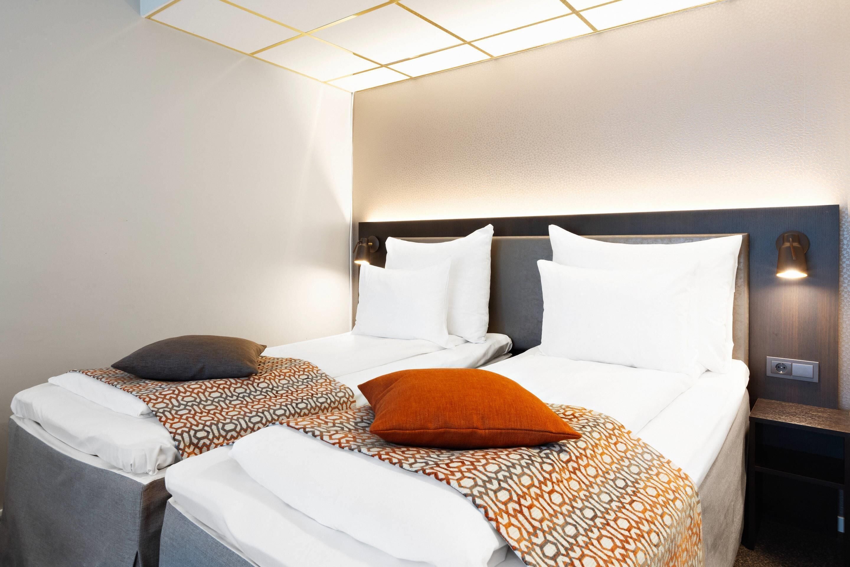Hotellihuone, jossa on kaksi sänkyä. Jokaisessa sängyssä on kuviollinen päiväpeite ja tyynyjä. Sängynpäädyssä on integroidut valot, ja seinälle on asennettu valaisin.