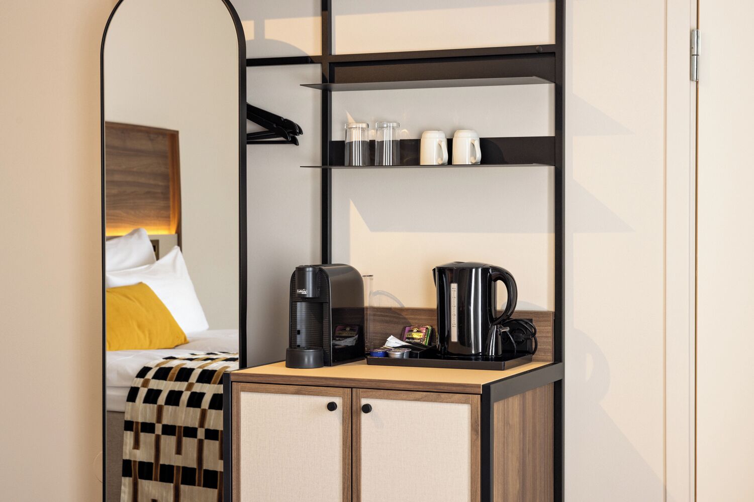 Habitación de hotel pequeña con cama, cafetera, hervidor y una pequeña estantería con tazas y una botella.