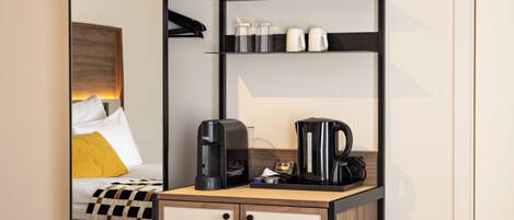 Habitación de hotel pequeña con cama, cafetera, hervidor y una pequeña estantería con tazas y una botella.