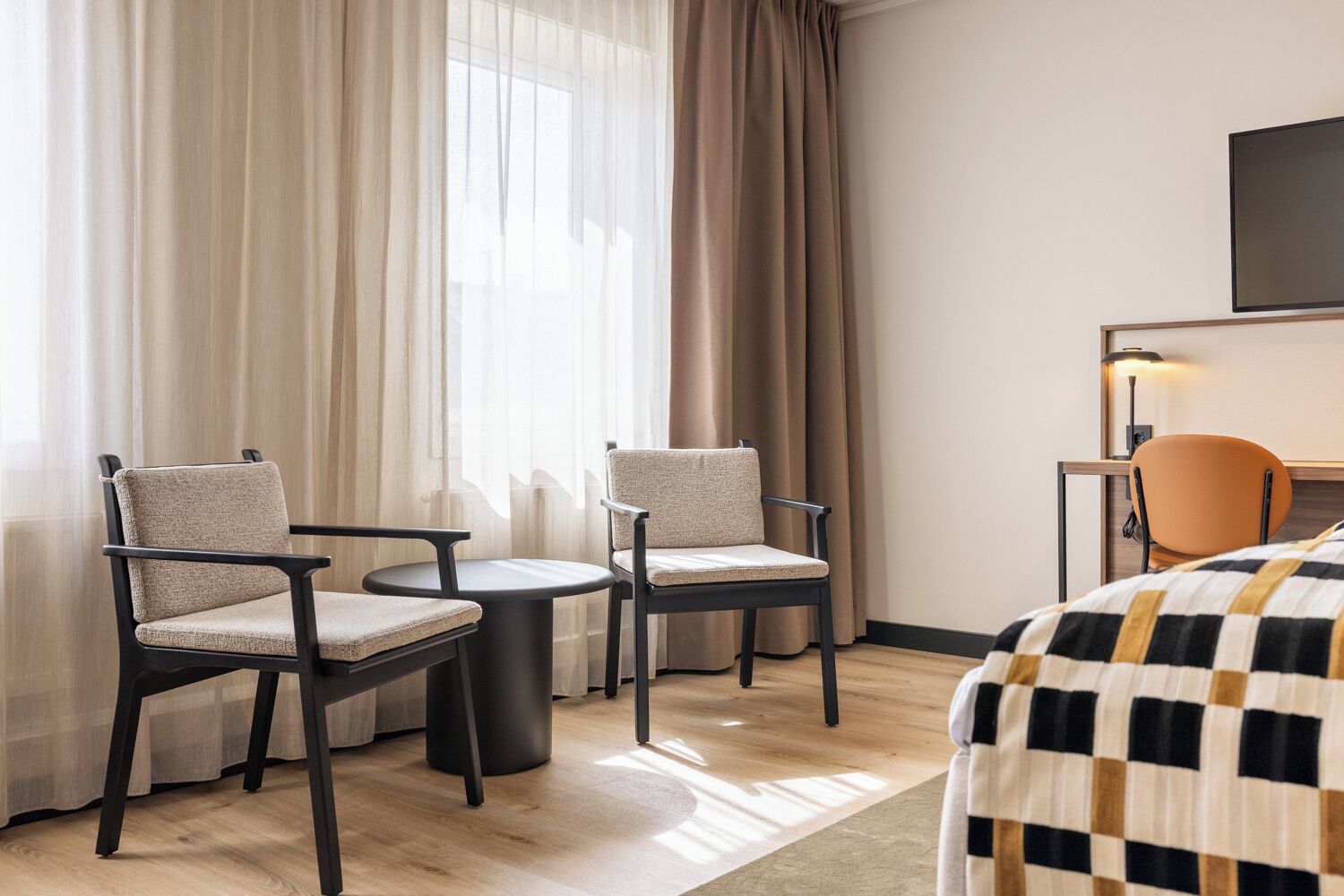 Habitación de hotel con cama, dos sillas, una mesita, un escritorio con lámpara y televisor en la pared.