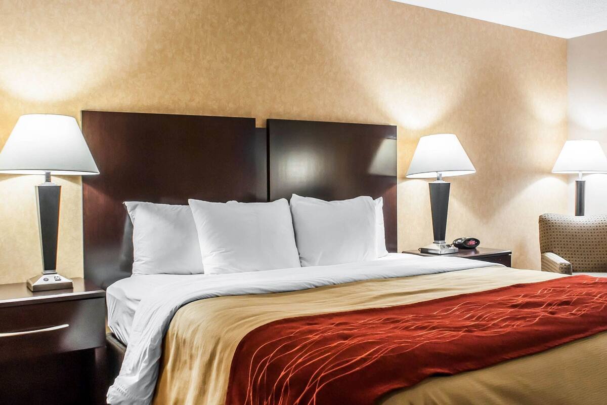 A hotel room with a large bed, two bedside tables with lamps, a red throw blanket, and a wooden headboard.