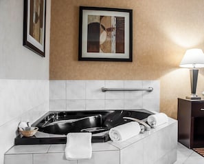 A modern bathroom with a black jacuzzi tub, white tiles, a framed artwork above the tub, and a bedside table with a lamp.