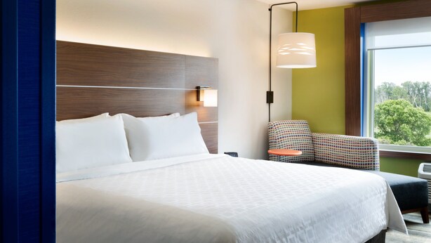 A hotel room with a large bed, a nightstand, a chair, a lamp, and a window with a view of trees. - Holiday Inn Express Canton by IHG (Canton)