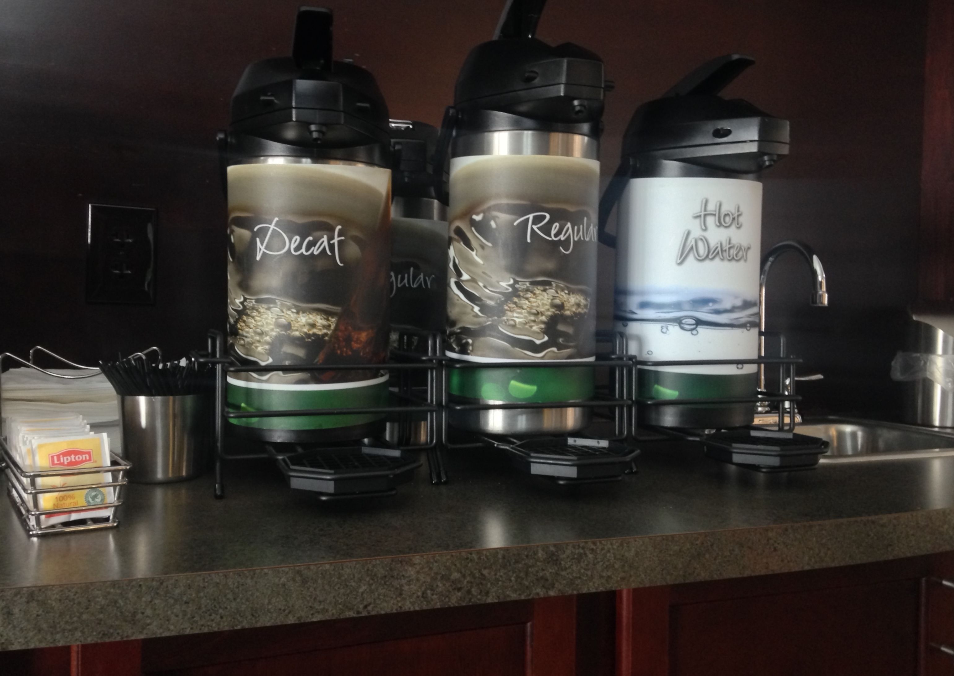 Una encimera de cocina con tres cafeteras etiquetadas 'Decaf', 'Regular' y 'Agua Caliente'.