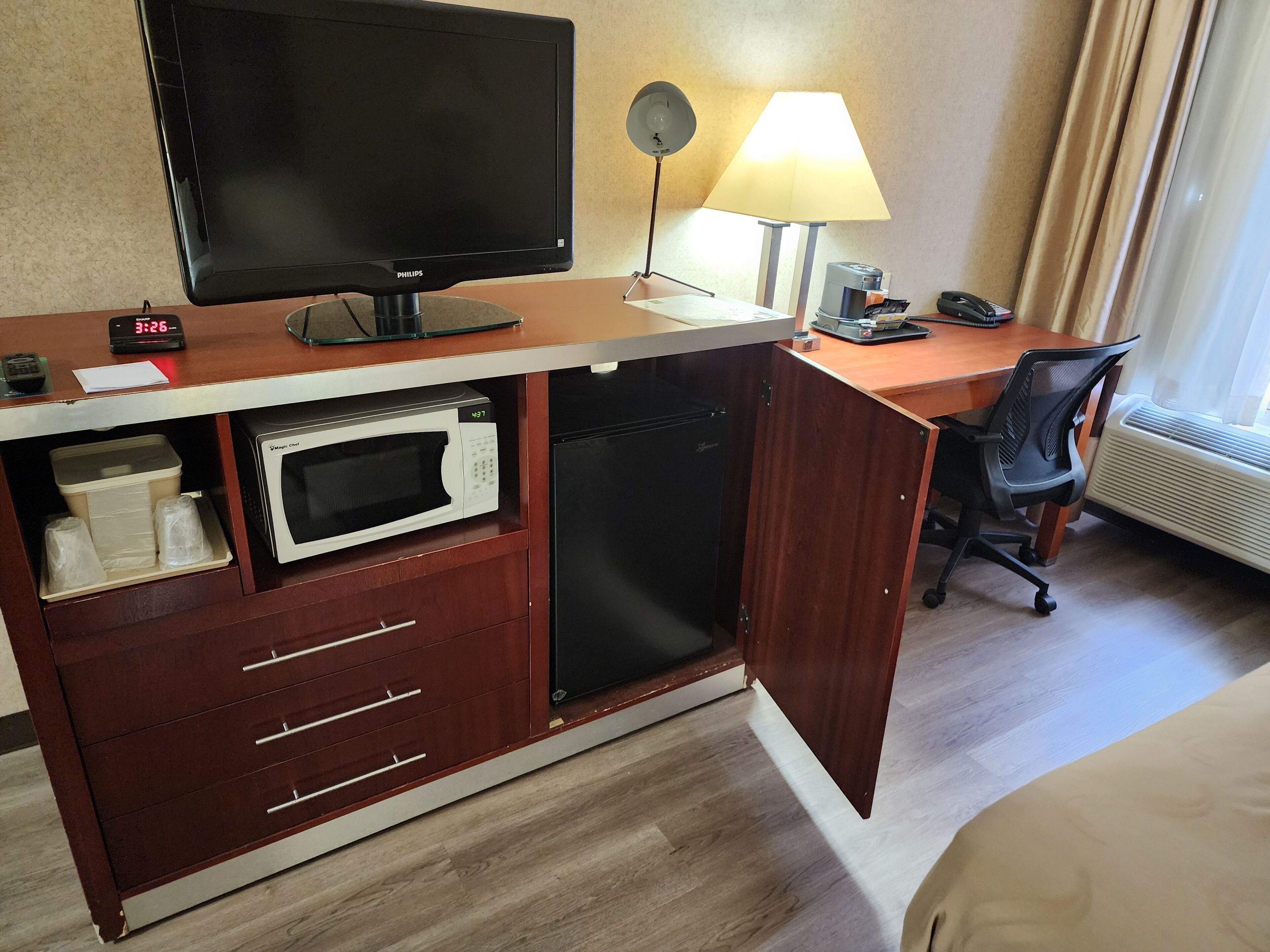 Ett hotellrum med ett skrivbord, en platt-TV, en mikrovågsugn, ett litet kylskåp, en stol, en lampa och en väckarklocka.