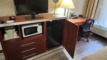 Ett hotellrum med ett skrivbord, en platt-TV, en mikrovågsugn, ett litet kylskåp, en stol, en lampa och en väckarklocka.