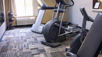 Un gimnasio con cintas de correr y equipo de ejercicio, piso alfombrado y una ventana con cortinas.