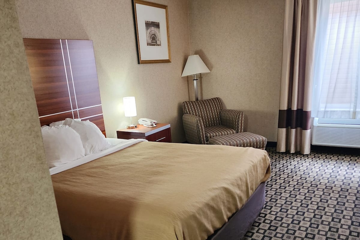 Een hotelkamer met een groot bed, een stoel, een bureau met een lamp, een ingelijst schilderij aan de muur en een raam met gordijnen.
