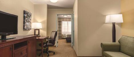 A hotel room with a television, desk, chair, couch, and a door leading to a bathroom.