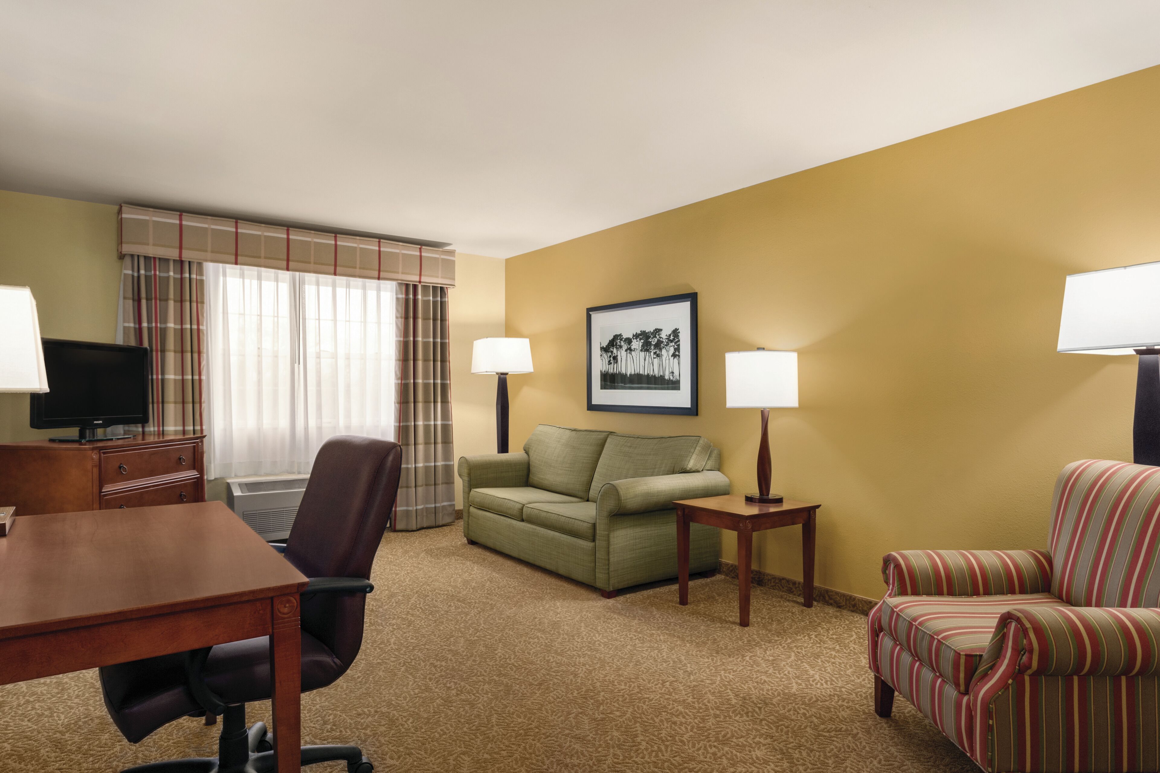 A hotel room with a desk, two chairs, a sofa, a television, and two lamps.