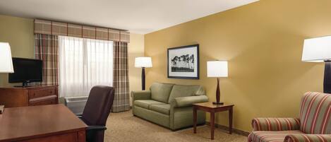 A hotel room with a desk, two chairs, a sofa, a television, and two lamps.