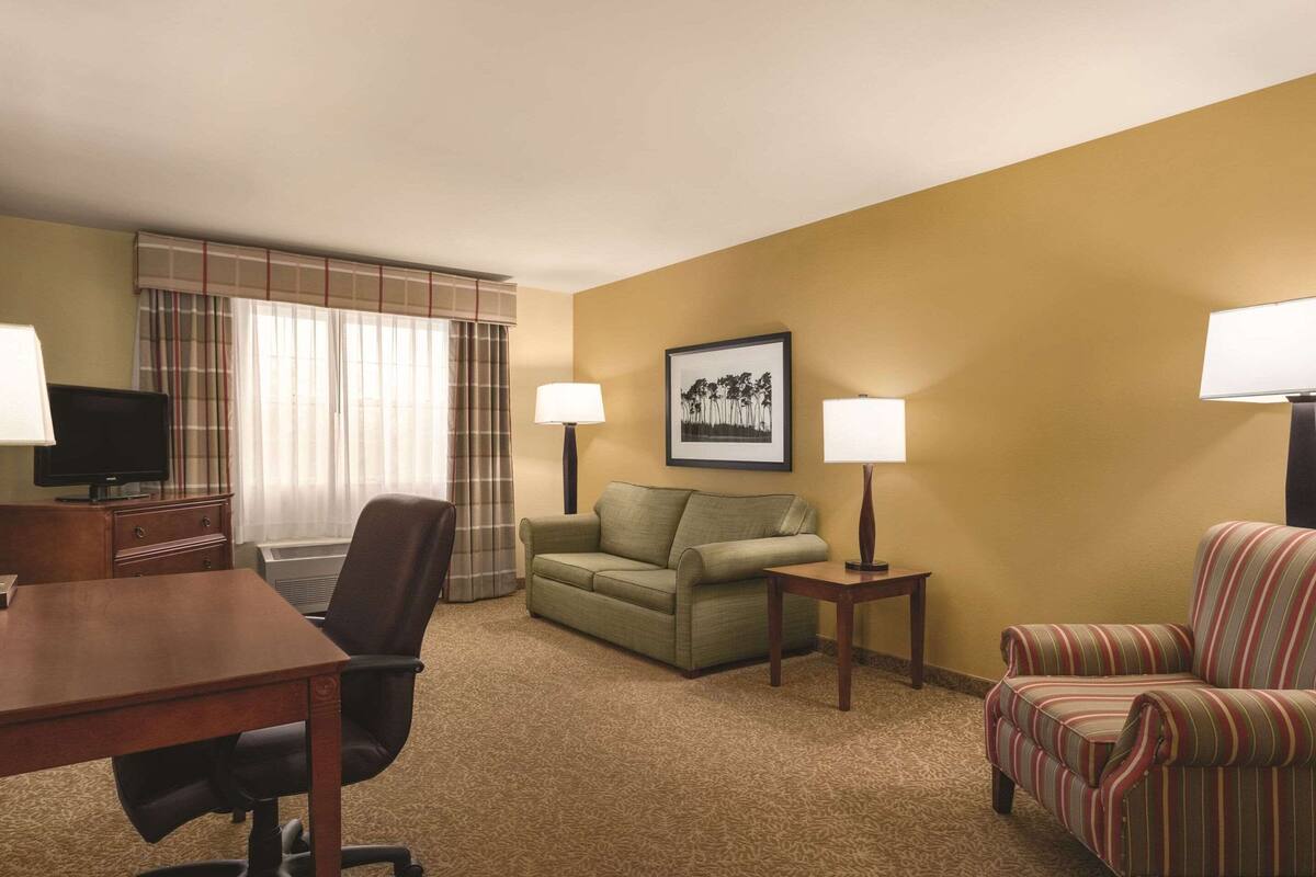 A hotel room with a wooden desk, a green sofa, a striped armchair, two lamps, a television, and a framed picture on the wall.