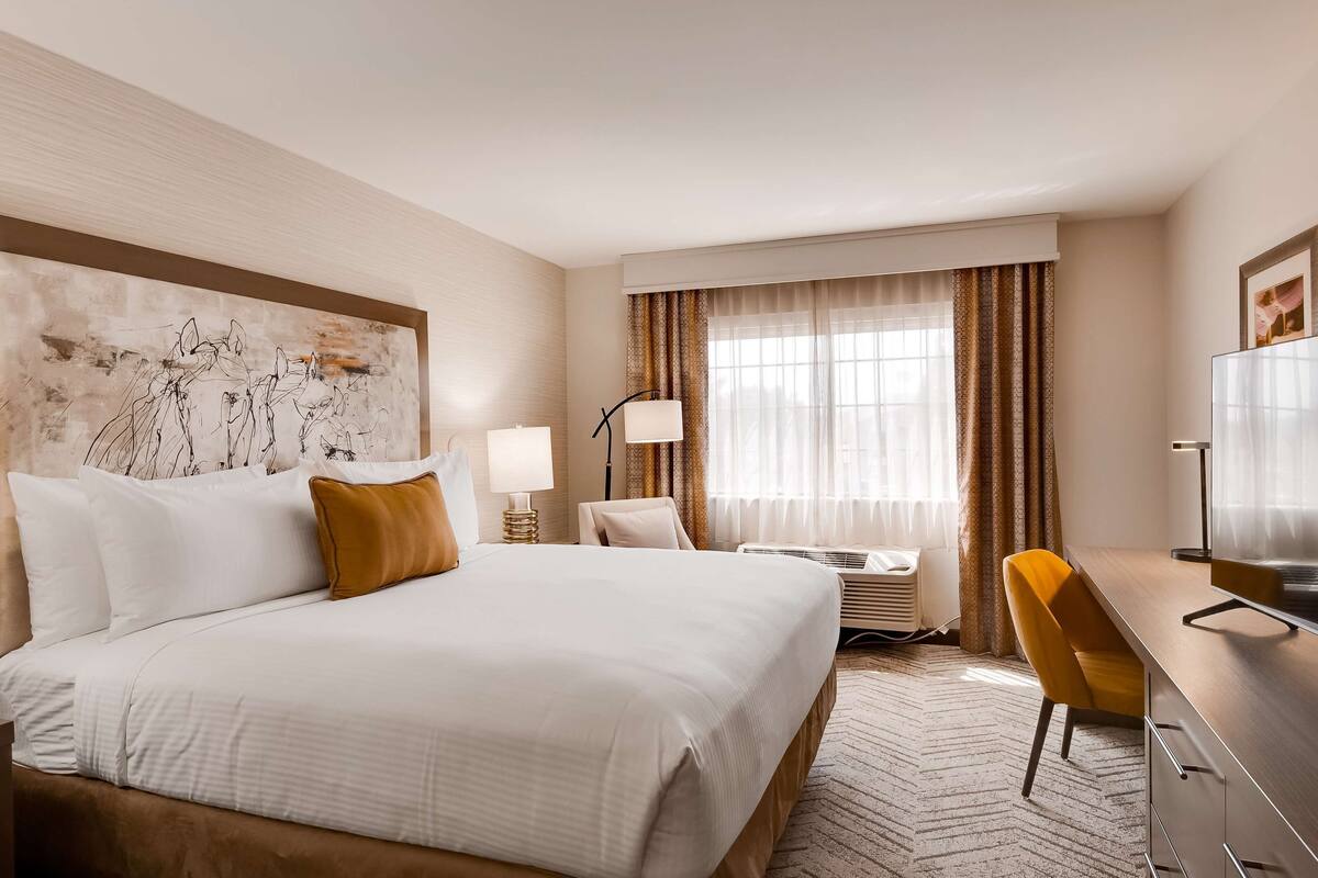 Une chambre d’hôtel avec un grand lit, un bureau avec une chaise, une télévision et une fenêtre avec des rideaux.