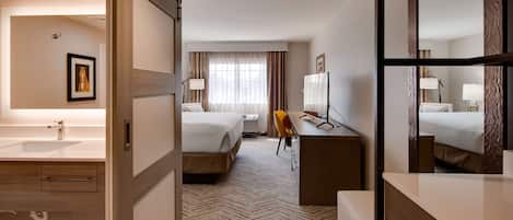 Una habitación de hotel con una cama grande, un escritorio con silla, una lámpara de escritorio, un espejo grande, un baño con lavabo y espejo, y una alfombra estampada.