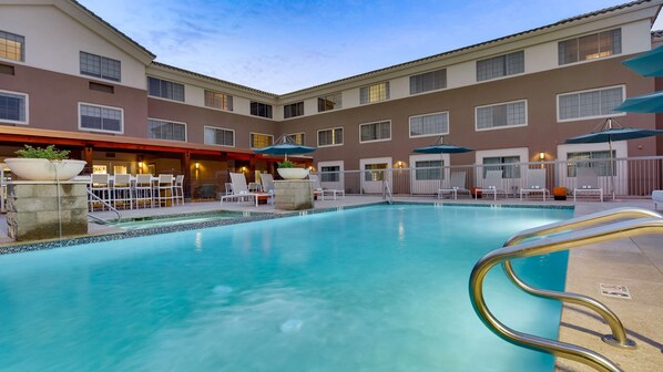 A hotel pool area with a clear blue pool, lounge chairs, umbrellas, and a bar area with seating.