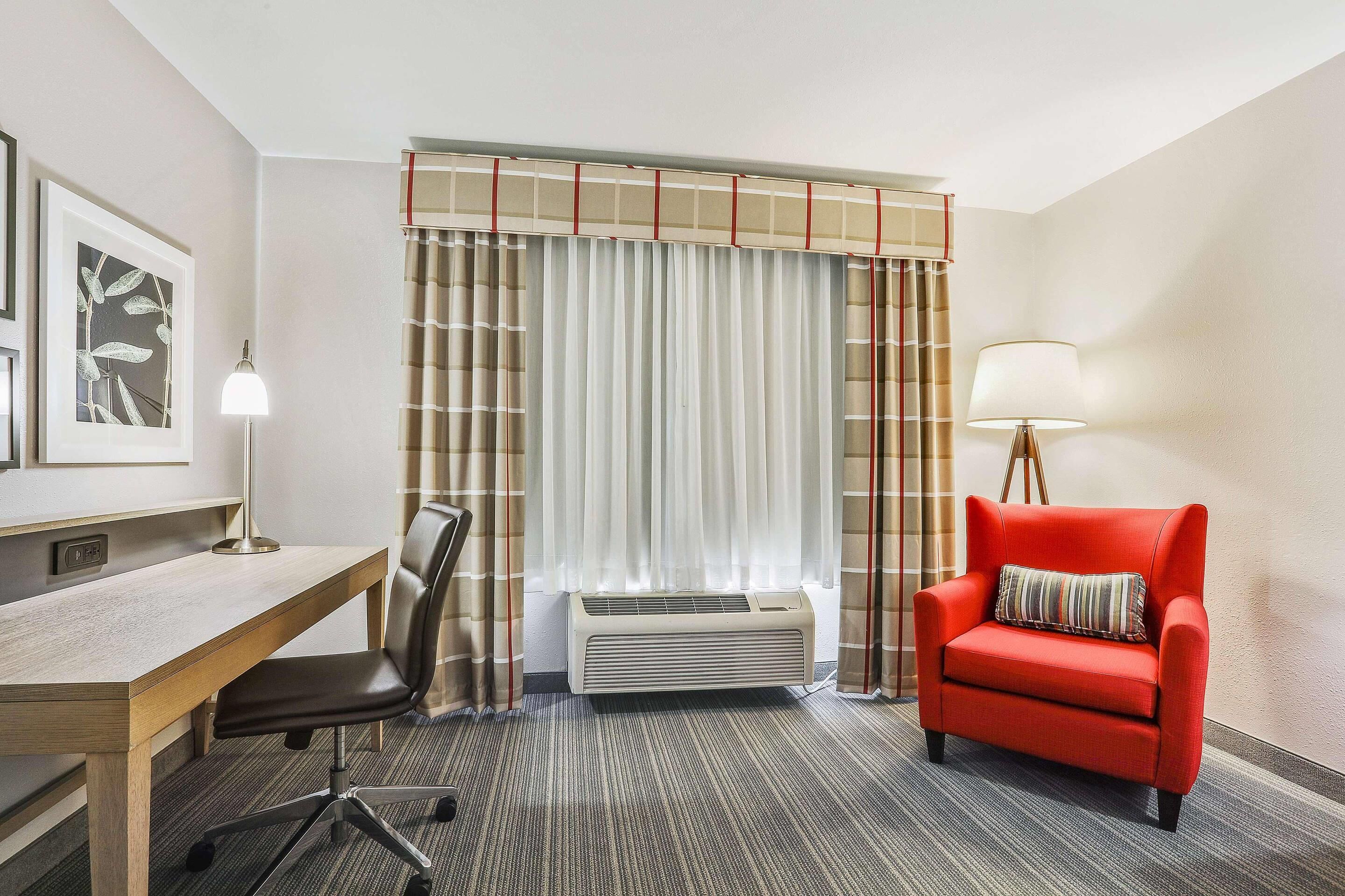 A hotel room with a desk, chair, red sofa, and a large window with curtains.