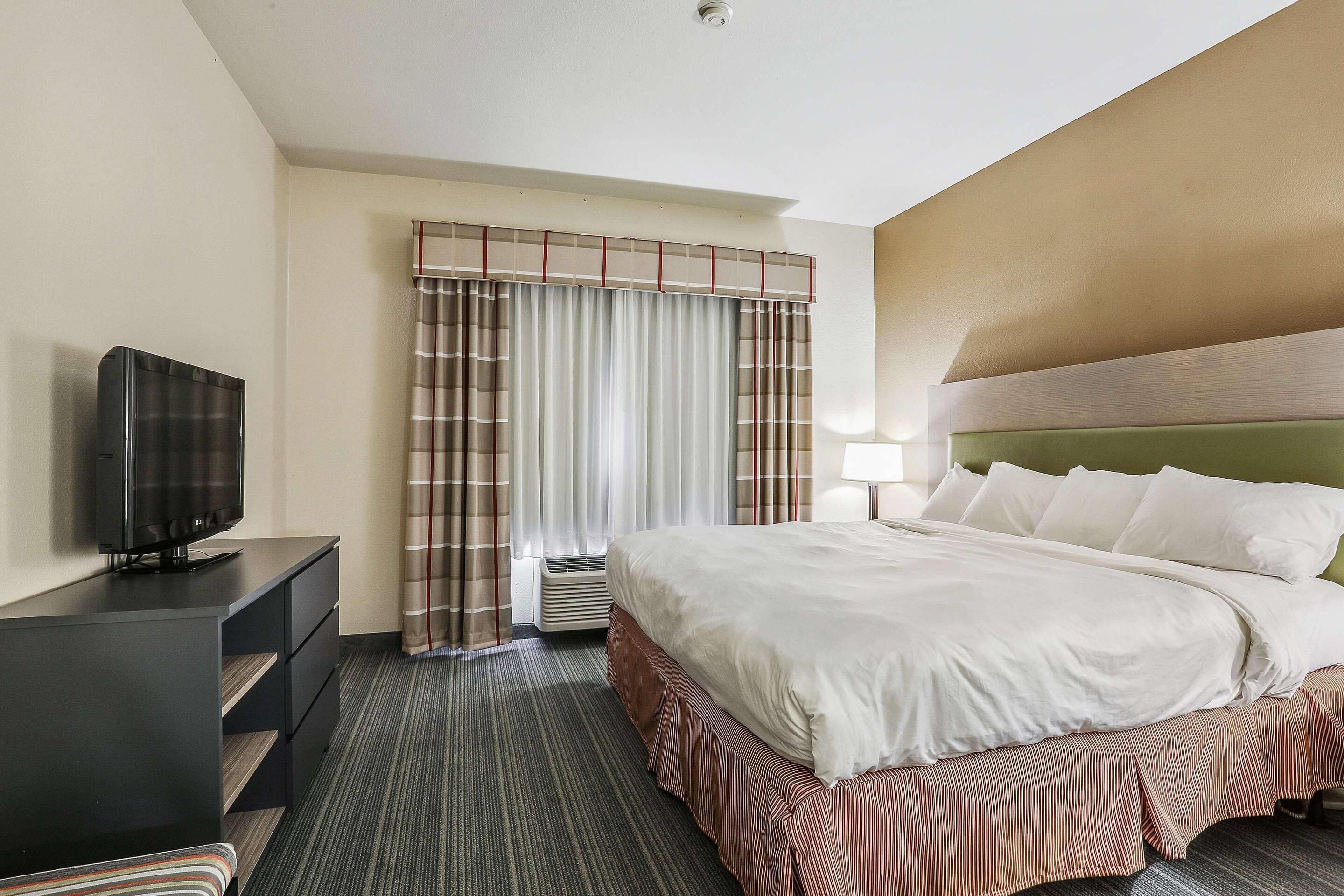 A hotel room with a bed, a TV on a stand, a desk, and a window with curtains.