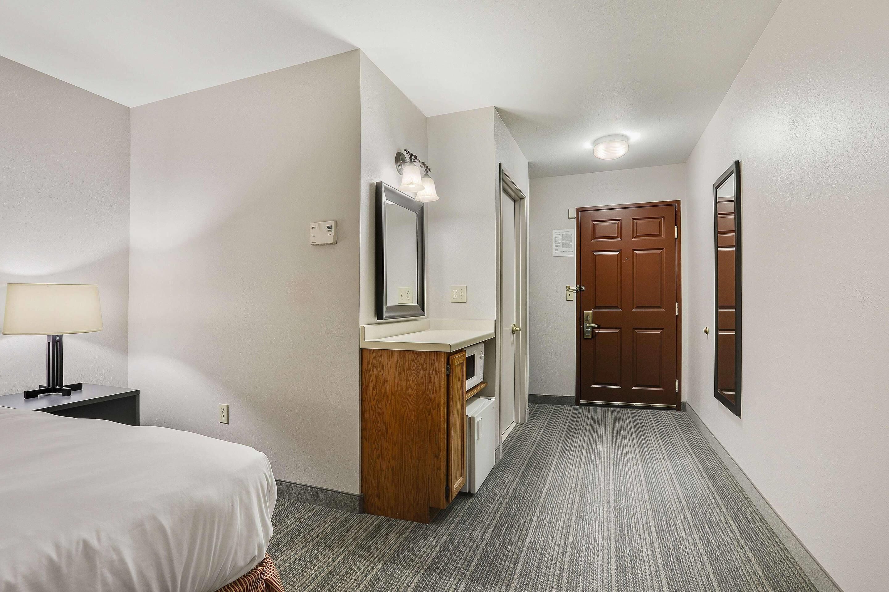 A hotel room with a bed, a vanity with a mirror, a door, and a carpeted hallway.