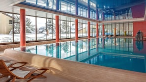 An indoor swimming pool with clear glass walls, wooden lounge chairs, and a view of snowy trees outside. - Dorint Blüemlisalp (Beatenberg)