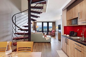 A modern kitchen with wooden cabinets, a built-in microwave, and a red backsplash. A spiral staircase leads to a balcony with a view of the mountains. - Dorint Blüemlisalp (Beatenberg)