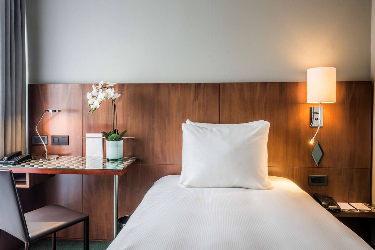 A hotel room with a bed, a desk with a chair, a telephone, a lamp, and a vase with flowers.