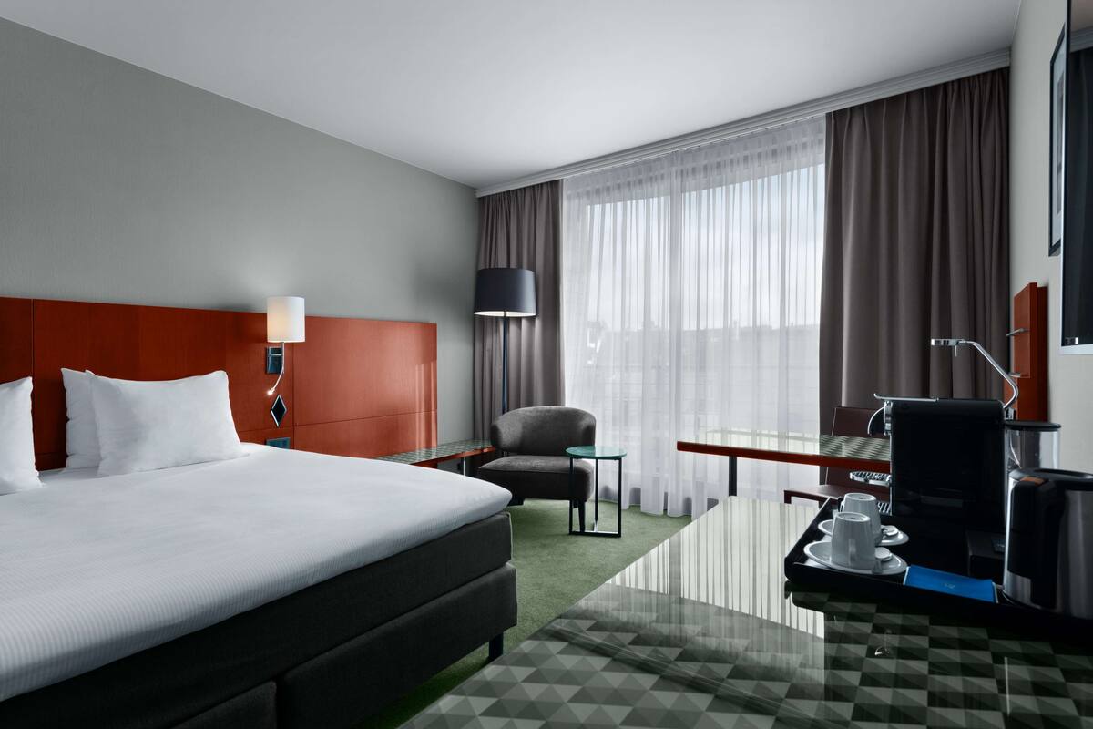 A hotel room with a large bed, a desk with a coffee maker, a chair, and a view of the city through large windows.