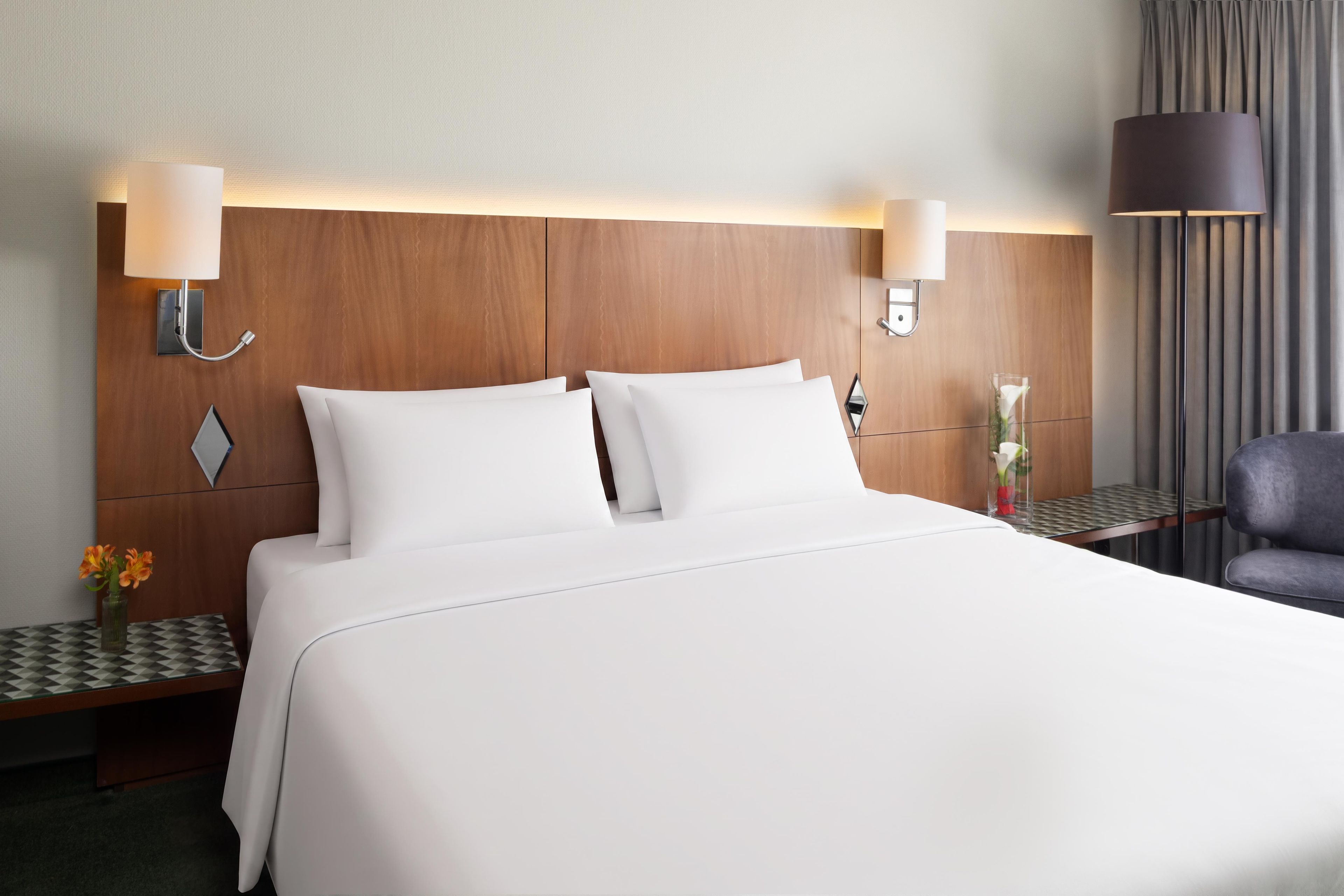 Una habitación de hotel con una cama grande, dos lámparas de noche, una mesita de noche con un florero y una silla.