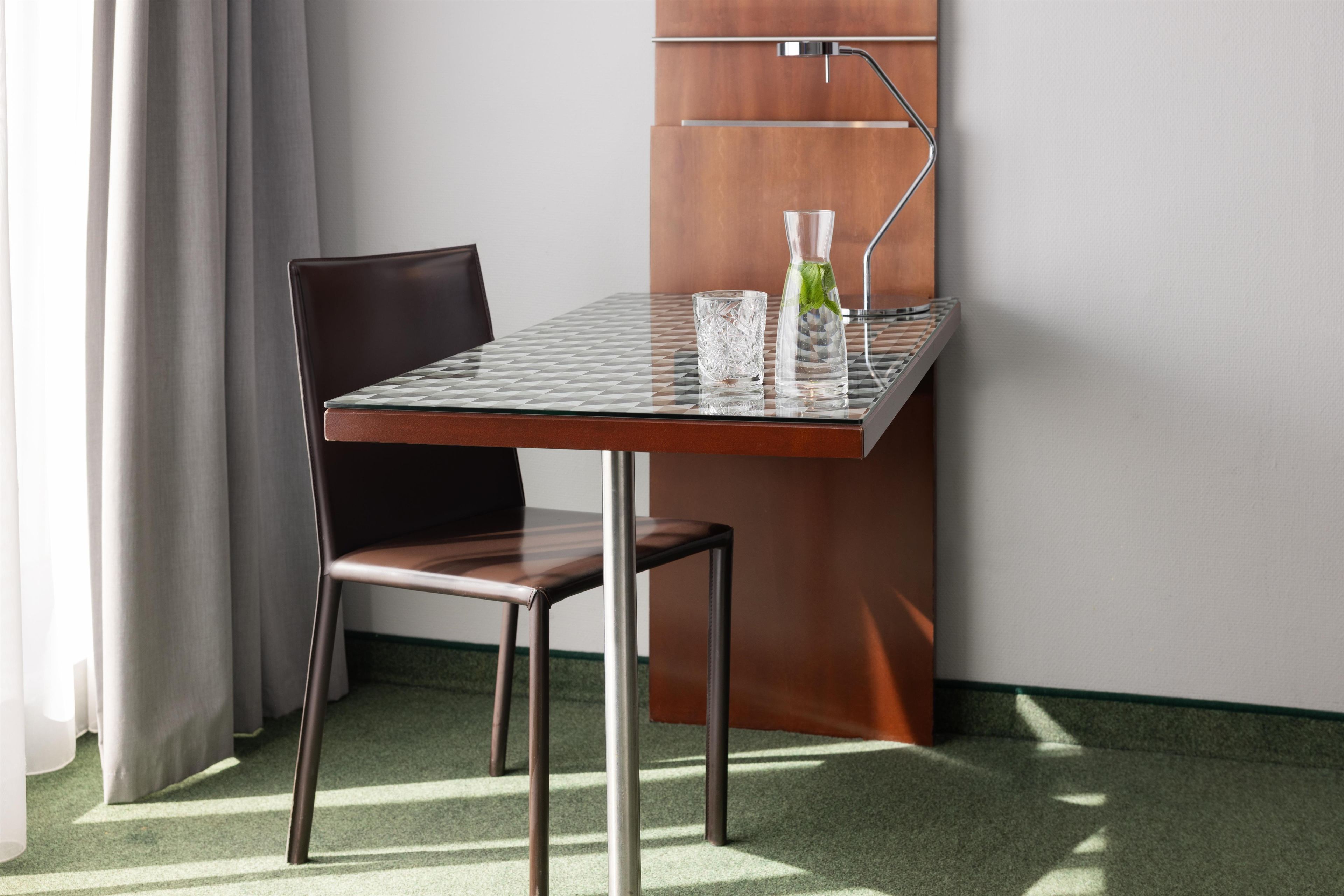 Een kleine, moderne kamer met een tafel met glazen blad, een stoel en een waterkan met een muntblaadje.