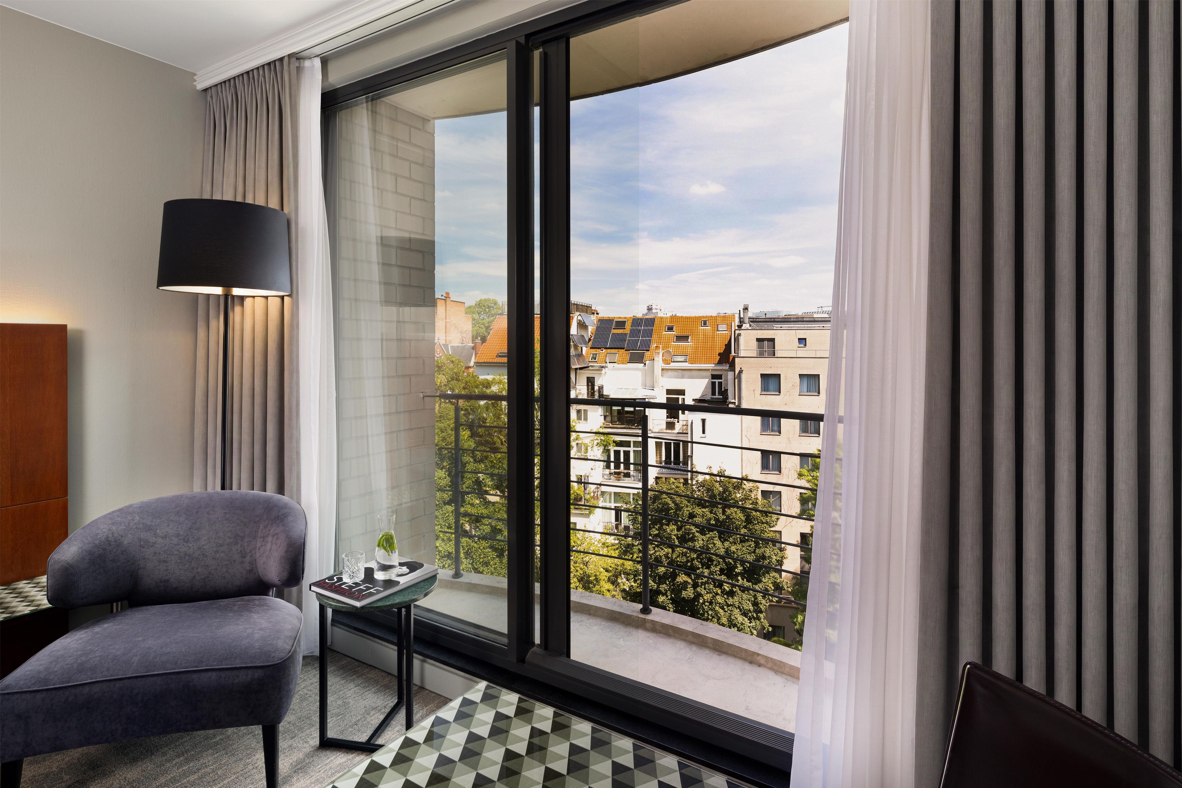 A modern hotel room with a large window, a grey armchair, a small table, and a floor lamp.